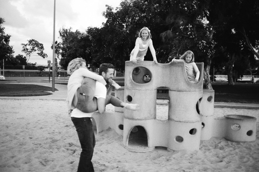 Wife and Husband wrestling as kids watch on, and laugh in park. ©William Bay Photographic Arts.